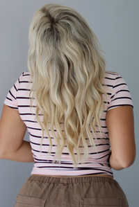 A woman shown from behind wearing a pink baby tee that has a burgundy and white striped design throughout, paired with mocha barrel denim, standing indoors against a white wall.