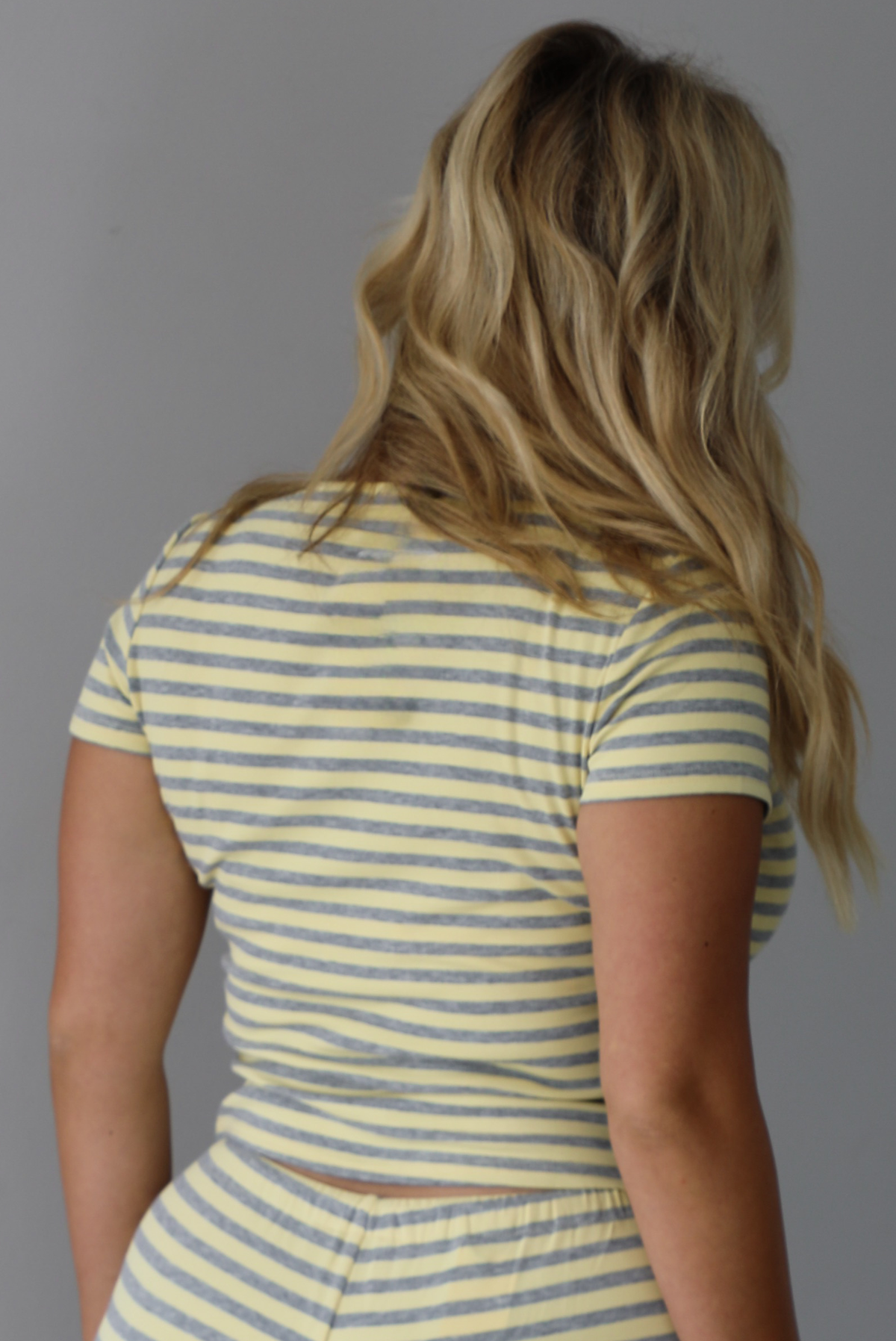 The back of a woman wearing a two piece set consisting of a collared, button-down, short sleeve top and high waisted, drawstring, wide leg pants, standing indoors against a white wall.