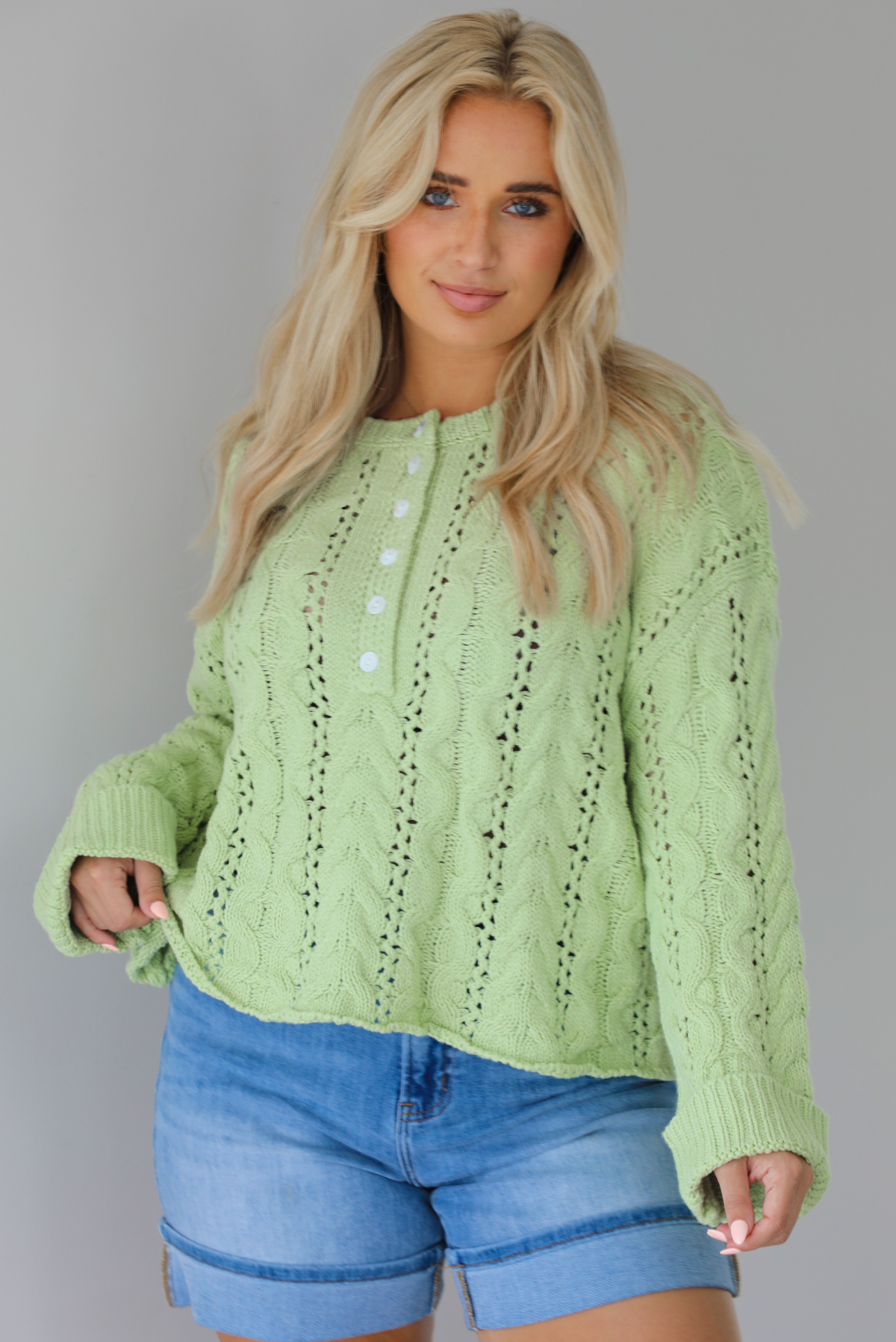 Woman wearing a green, long sleeved sweater with a loose knit design, functional button closures in the front, cuffed sleeves, and a raw, unfinished hem with medium wash denim shorts indoors against a white wall.