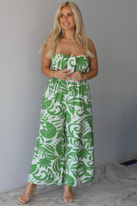 Woman wearing a strapless jumpsuit with green leaf pattern indoors against a white wall with white marble flooring. 