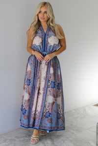 Woman wearing a blue and multi-colored halter tie maxi dress that has a long, flowy skirt with a split hem detail. The photo is taken indoors against a white wall with white marble flooring. 