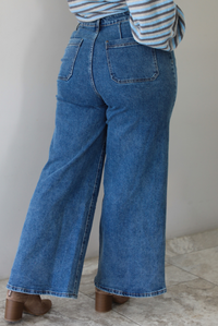 A woman shown from behind wearing blue wide-leg denim jeans with back pockets, paired with a striped long-sleeve top and tan pointed-toe booties, standing indoors against a white wall with white marble flooring. 