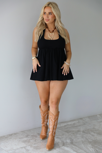 Woman wearing a black romper that looks like a dress, with a halter tie neckline, baby-doll cut, and flowy skirt indoors against a white wall with white marble flooring. 