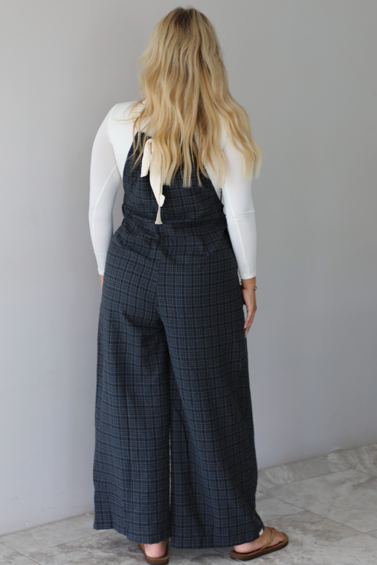 A woman shown from behind modeling a white long-sleeve top and dark gray plaid wide-leg overalls, standing indoors against a white wall with white marble flooring. 