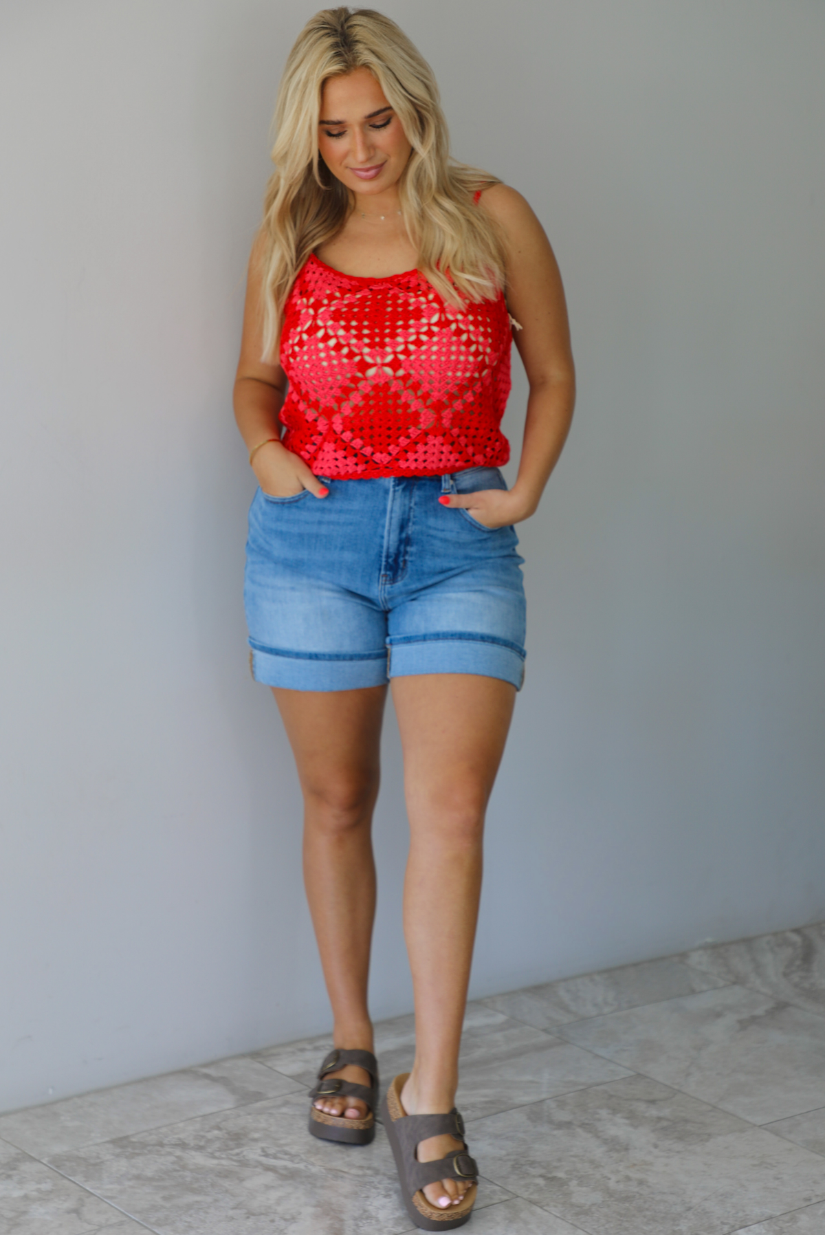 Woman wearing a pink and red loose knit crochet cropped top with light wash denim shorts indoors against a white wall with white marble flooring. 