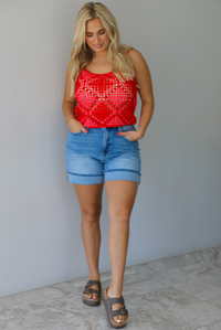 Woman wearing a pink and red loose knit crochet cropped top with light wash denim shorts indoors against a white wall with white marble flooring. 