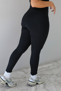 Woman wearing a fitted two piece black set featuring a short sleeved top and fitted leggings. The photo shows a closeup of the back of the pants, which is paired with white ankle socks and silver sneakers.  The photo is taken indoors against a white wall and white marble flooring. 