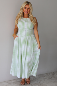 A woman wearing a light green and white gingham maxi dress with a drop waist bodice and side pockets, standing indoors against a white wall with white marble flooring. 