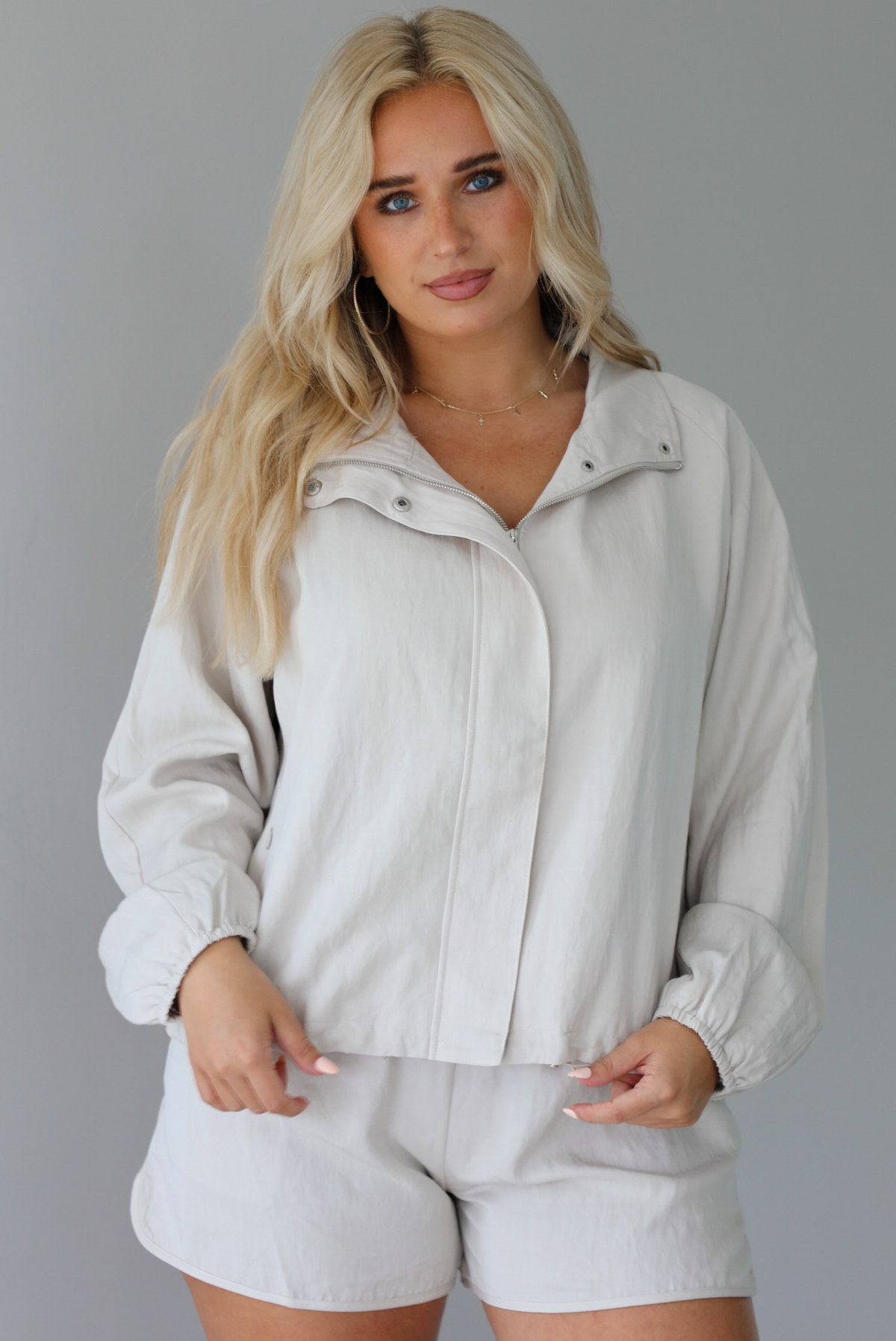 Woman wearing a stone colored two piece set featuring a zip up jacket and shorts with a navy tank tucked into the shorts indoors against a white wall. The jacket is shows zipped up. 