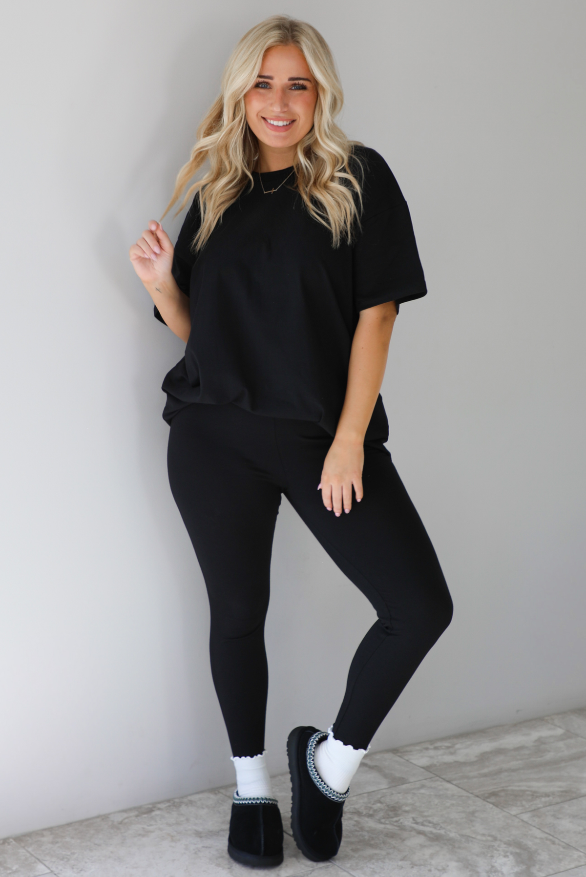 Woman wearing a black two piece set featuring an oversized t-shirt and fitted leggings. The set is paired with high white ankle socks and black slip on shoes. The photo is taken indoors against a white wall with white marble flooring. 