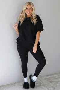 Woman wearing a black two piece set featuring an oversized t-shirt and fitted leggings. The set is paired with high white ankle socks and black slip on shoes. The photo is taken indoors against a white wall with white marble flooring. 