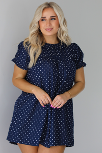 Woman wearing a navy baby-doll cut dress with a white polkadot print pattern throughout. The dress has puffed short sleeves and functional pockets on either side. The photo is taken indoors against a white wall.