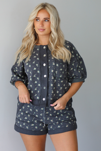 Woman wearing a charcoal with a multi-colored floral two piece set featuring an oversized top with puffed sleeves and shorts indoors against a white wall. The photo shows a close up of the set, showing the functional button closures on the front of the top and the elastic and drawstring waistline on the shorts. The top is untucked from the shorts showing its full length.