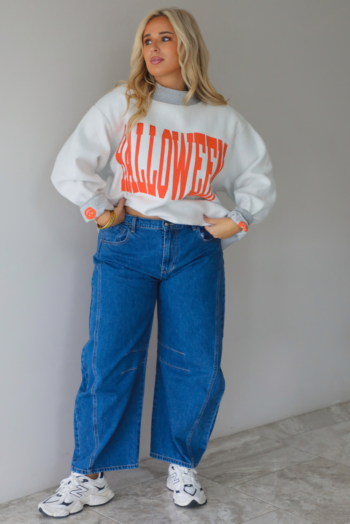 A woman wearing a light grey and dark grey trim oversized sweatshirt that says "HALLOWEEN" in collegiate font on the front, with jack-o-lantern details on the rolled up sleeves, paired with dark wash denim jeans, standing indoors against a white wall with white marble flooring. 