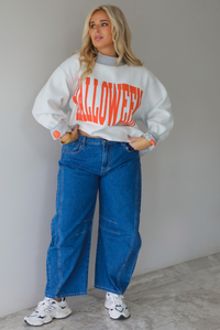 A woman wearing a light grey and dark grey trim oversized sweatshirt that says "HALLOWEEN" in collegiate font on the front, with jack-o-lantern details on the rolled up sleeves, paired with dark wash denim jeans, standing indoors against a white wall with white marble flooring. 