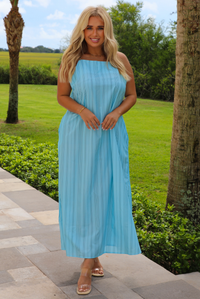 A woman wearing a light blue sleeveless maxi dress with vertical grey stripes, standing outdoors on a stone pathway with a green lawn and trees in the background.