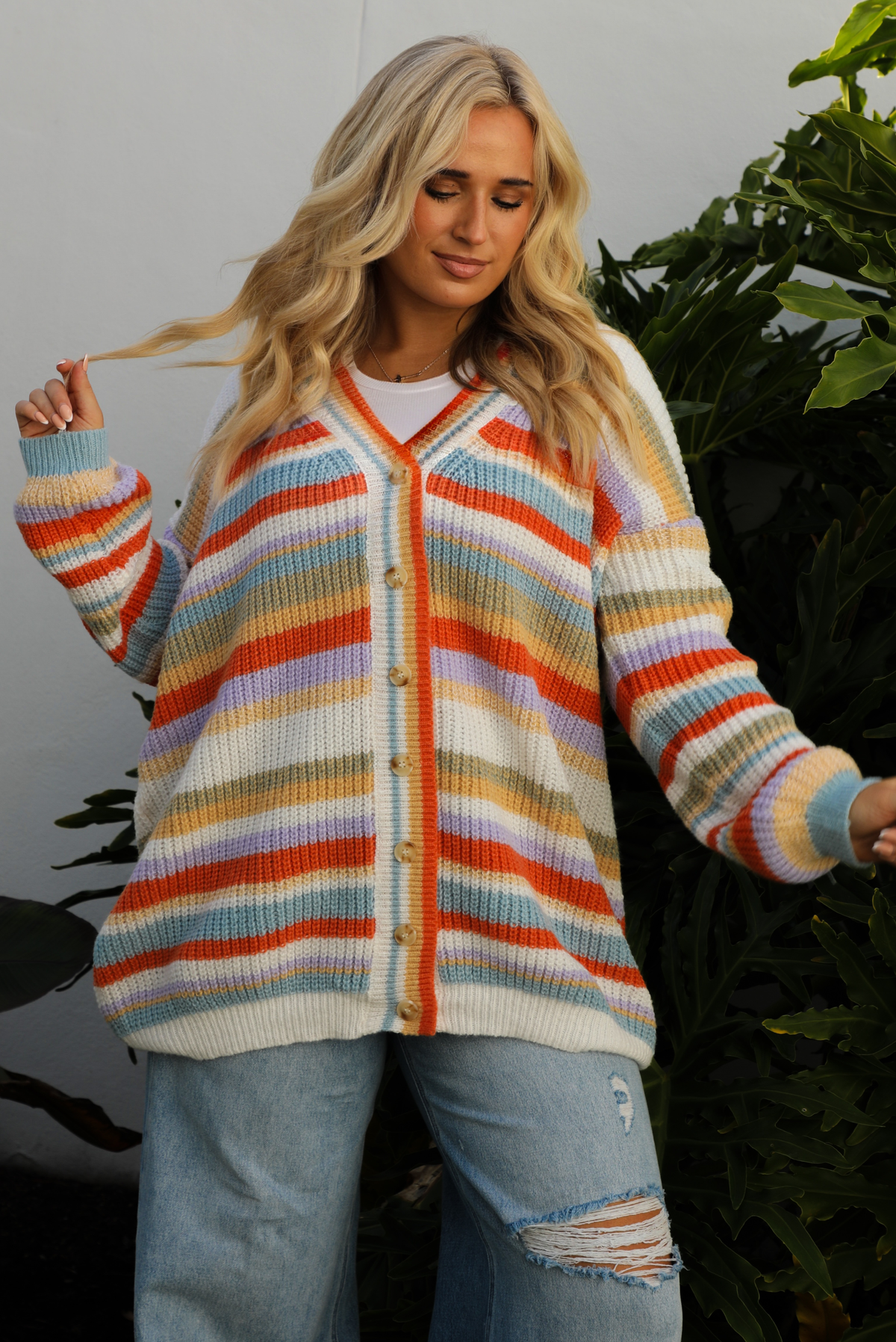 Woman model wearing an orange, lavender, yellow, muted green, blue, and white striped oversized cardigan with functional button closures in the front and an oversized fit with a light wash flared hem denim. The photo is taken outdoors against a white wall and greenery in the back. The photo showcases a close up of the cardigan and shows the full length, which falls to the upper thigh on our 5'7" model.