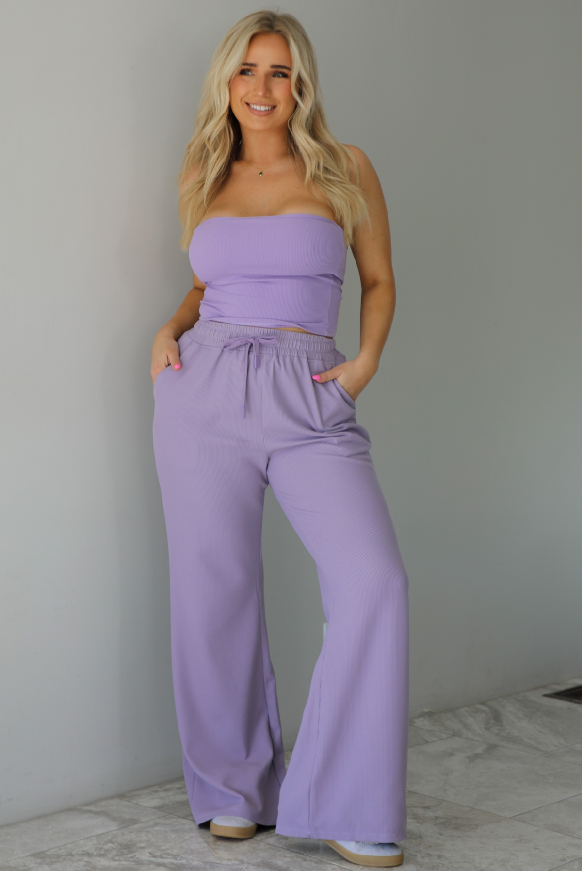 Woman wearing a two piece lavender set featuring a strapless, fitted tube top, and high waisted, flared hem pants. The photo is taken indoors against a white wall with white marble flooring.