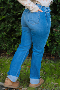 The photo shows a close up of the back of a pair of medium wash denim that have a mid-rise waistline, functional front and back pockets, a button and zipper closure in the front, slightly distressed details on the hips the bottom of the left thigh, and a cuffed hem. The photo shows the model with a a hand in the back right pocket, and is taken outdoors with greenery in the back. 