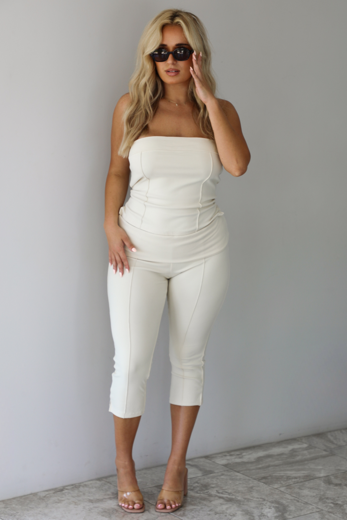 Woman wearing a cream two piece set featuring a strapless tube top and capri pants that fold over at the waist indoors against a white wall with white marble flooring. 