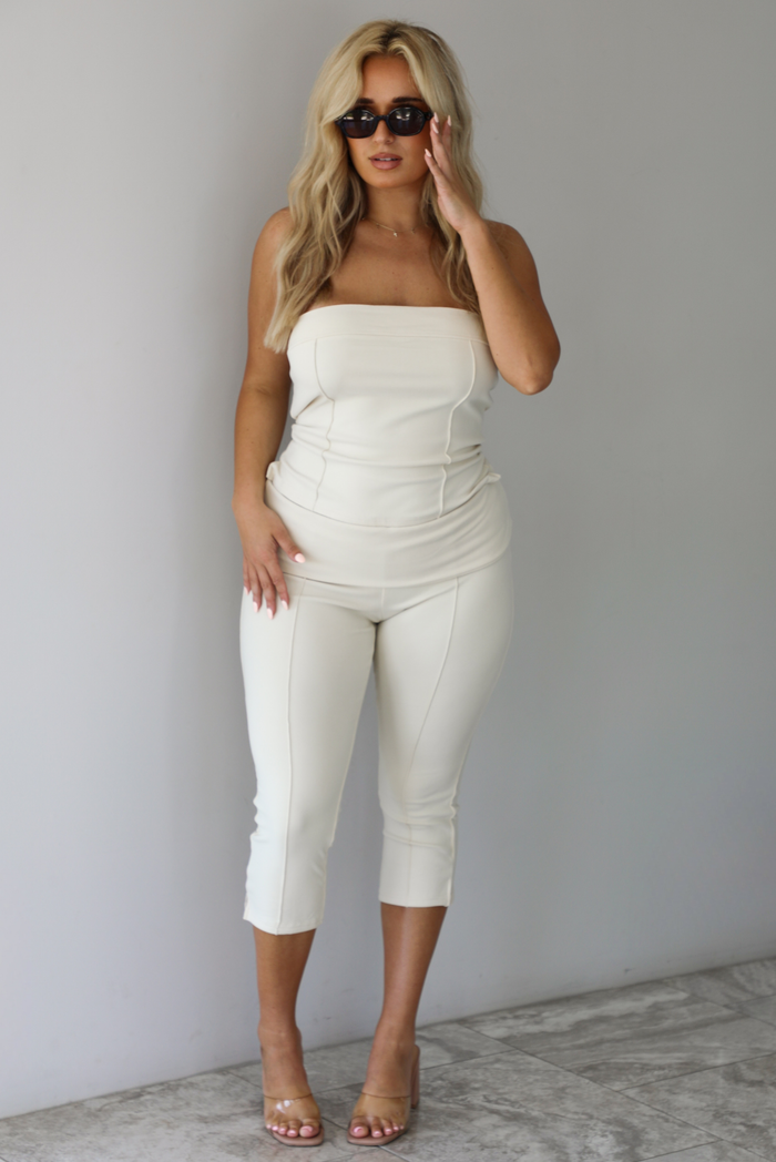 Woman wearing a cream two piece set featuring a strapless tube top and capri pants that fold over at the waist indoors against a white wall with white marble flooring. 