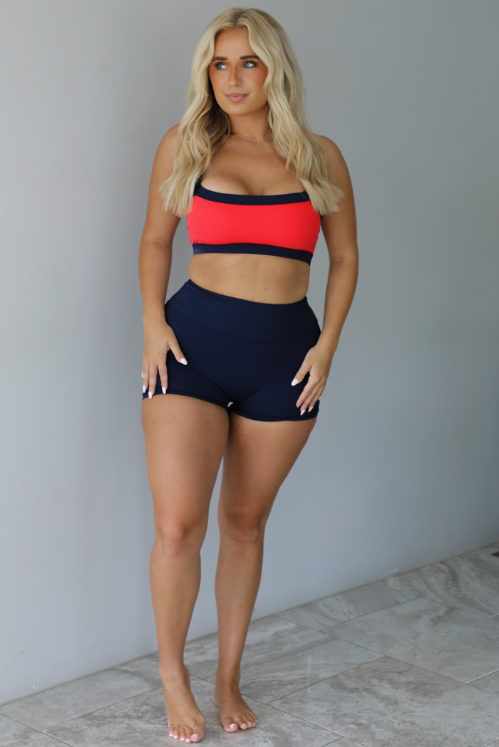 Woman wearing. navy and red sports bra with navy high waisted shorts indoors against a white wall with white marble flooring. 