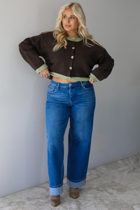 A woman wearing a dark brown with green trim cardigan fully buttoned up paired with dark wash, cuffed hem denim jeans, standing indoors against a white wall with white marble flooring. 