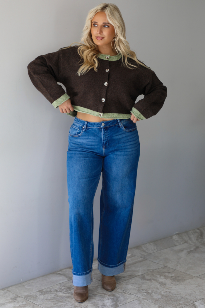 A woman wearing a dark brown with green trim cardigan fully buttoned up paired with dark wash, cuffed hem denim jeans, standing indoors against a white wall with white marble flooring. 