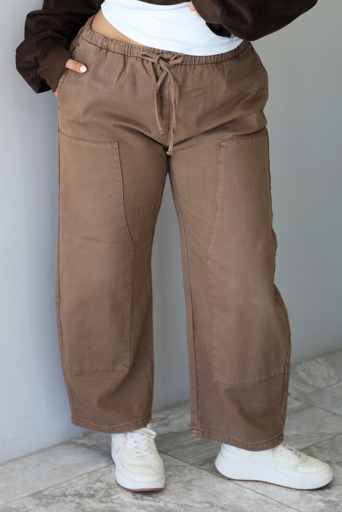 A closeup of a woman wearing mocha mid-to-low rise denim pants with a drawstring and elastic waistline and pockets on either side, paired with a white tank and dark brown suede jacket and cream sneakers, standing indoors against a white wall with white marble flooring. 