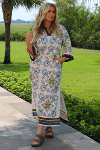 A woman wearing a bohemian-style printed kaftan dress with blue, green, and brown paisley patterns on a cream background, standing outdoors on a stone pathway with a lush green lawn and palm trees in the background.