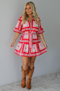 A woman modeling a red and white patterned short-sleeve mini dress with a tie waist, paired with brown cowboy boots, standing indoors against a white wall with white marble flooring. 
