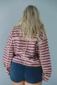Woman wearing a pink, burgundy, lilac, and sand colored jacket with a charcoal colored two-piece set featuring a cropped sports bra and high waisted short biker shorts. The photo shows the back of the jacket. The photo is taken indoors against a white wall.