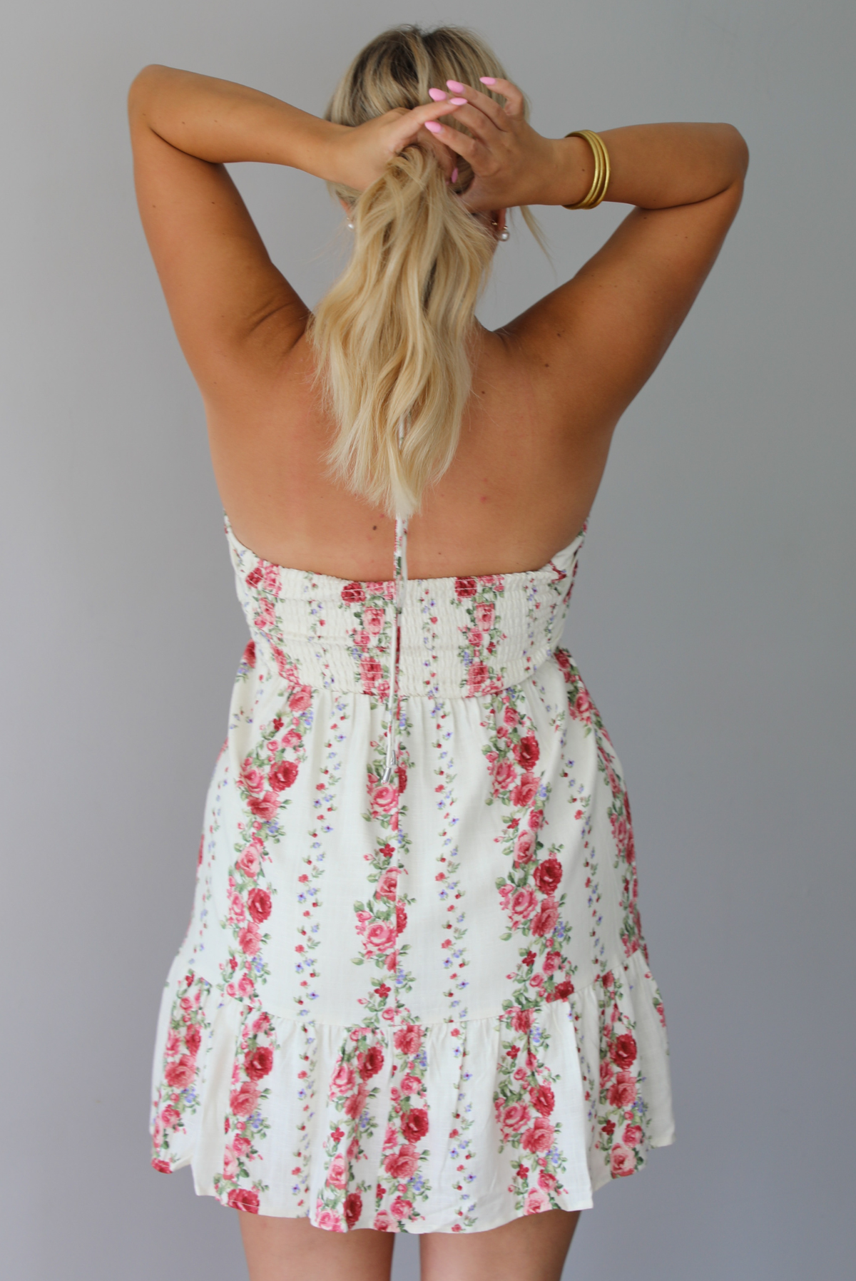 A woman wearing a white halter mini dress with a red and pink floral print, standing indoors against a white wall. The photo shows the back of the dress, which has a smocked detail.