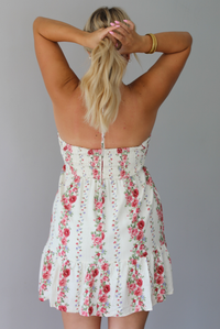 A woman wearing a white halter mini dress with a red and pink floral print, standing indoors against a white wall. The photo shows the back of the dress, which has a smocked detail.