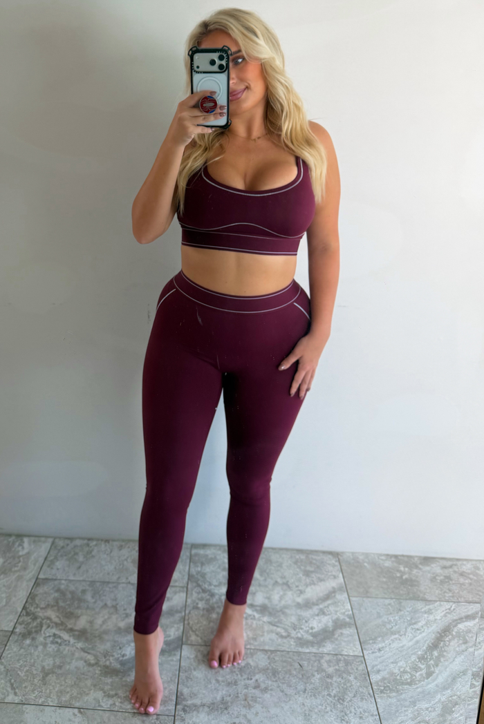 A woman wearing a maroon matching two piece set featuring a sports bra and high waisted leggings with white stitching details, taking a mirror selfie indoors against a white wall with white marble flooring. 