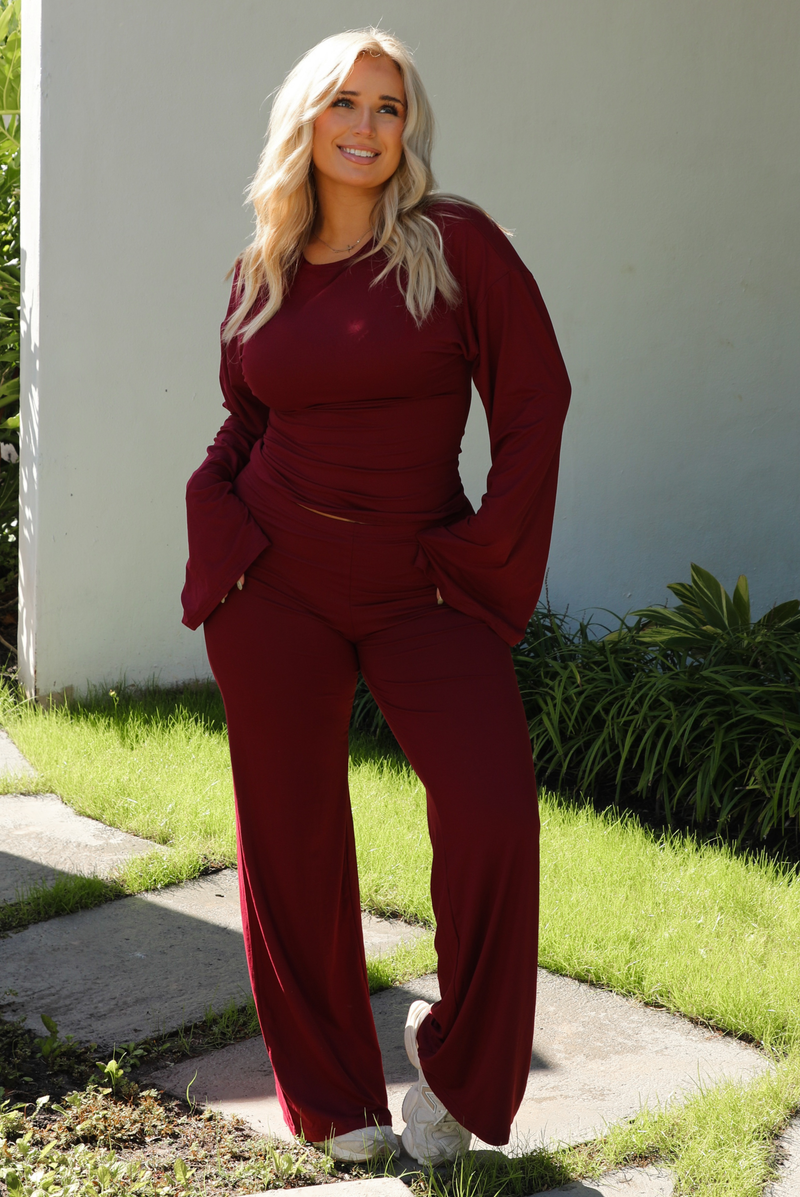 Woman model wearing a burgundy wine colored two piece set outdoors. The set features a slightly cropped, long sleeved top that has flared sleeves and a boat neckline. The pants are a mid-rise, fitted on the thighs, and have a straight cut design at the bottom. It's super soft, lightweight, and stretchy. 