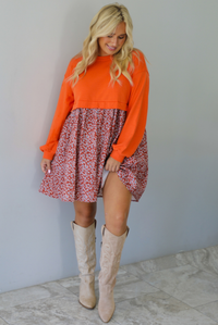 A woman wearing a block style oversized, flowy dress featuring a solid orange bodice and sleeves with a multi-colored floral skirt, paired with tall sand colored boots, standing indoors against a white wall with white marble flooring. 