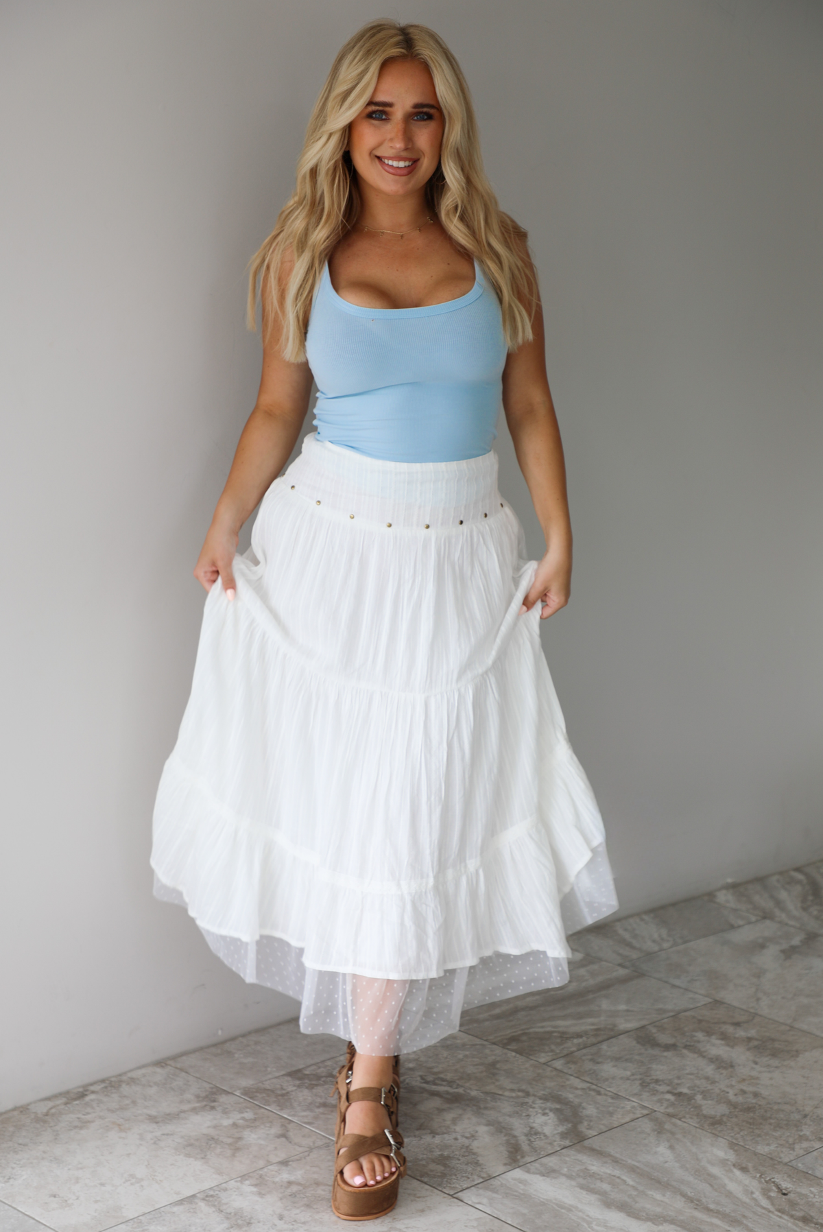 Woman wearing a blue tank and high waisted white maxi-length skirt indoors against a white wall with white marble flooring. 
