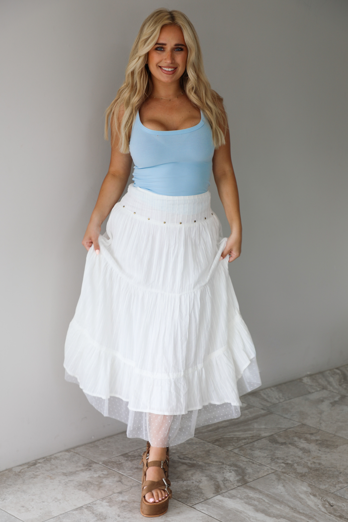 Woman wearing a blue tank and high waisted white maxi-length skirt indoors against a white wall with white marble flooring. 