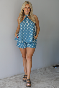 Woman wearing denim blue with white polka dot halter tie top and high waisted shorts indoors against a white wall with white marble flooring. 