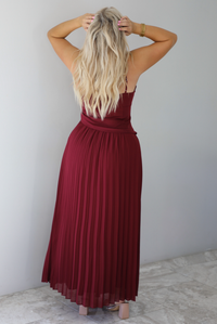 A woman shown from behind wearing a wine colored formal maxi dress with a fitted, drop waist design with a crimped floral maxi skirt, standing indoors against a white wall with white marble flooring. 