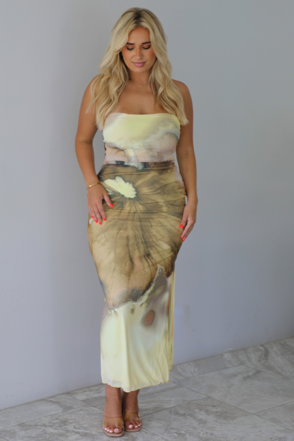 Woman wearing a yellow and brown floral print, strapless, fitted midi dress indoors against a white wall with white marble flooring. 