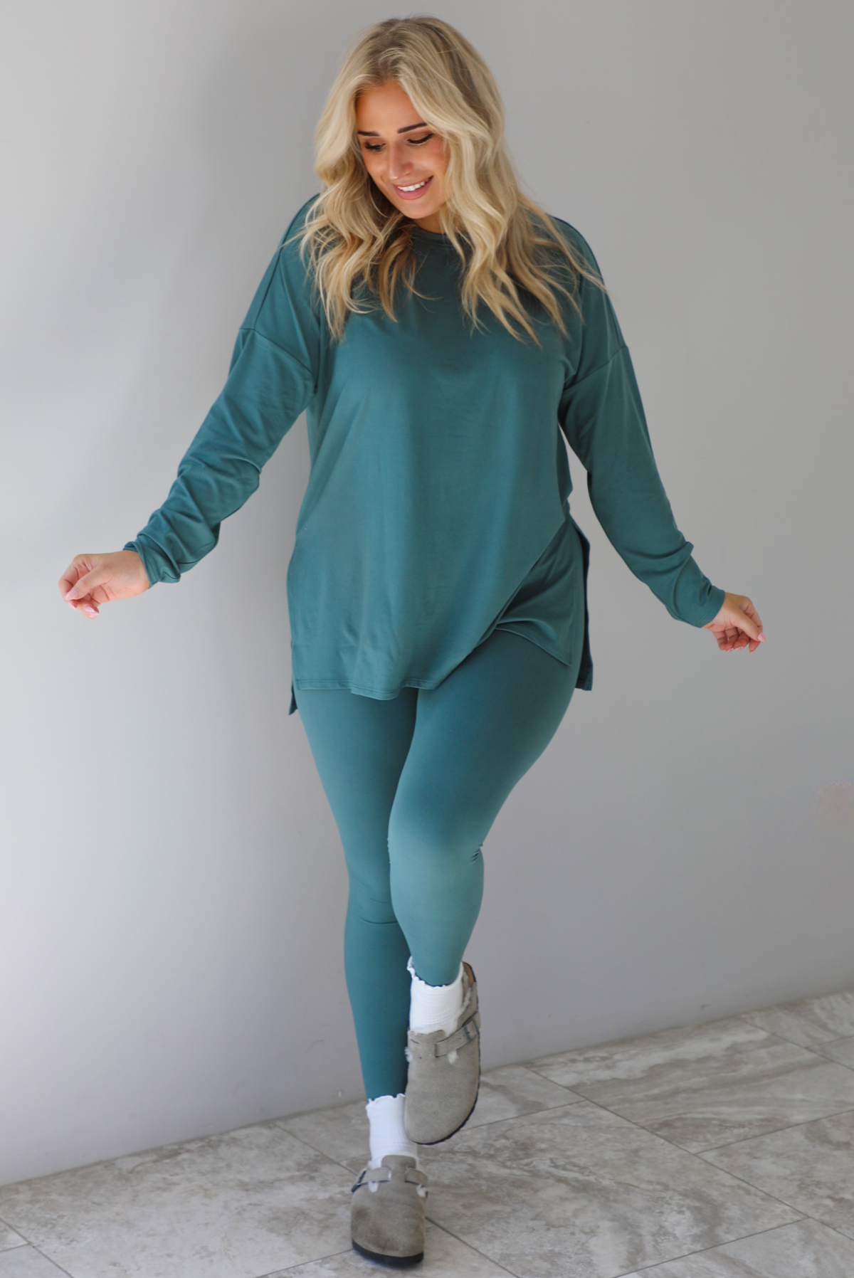 Woman wearing a teal two piece set that features an oversized long sleeved top and fitted leggings. The set is paired with high ankle socks and grey slide on shoes. The photo is taken indoors against a white wall with white marble flooring.