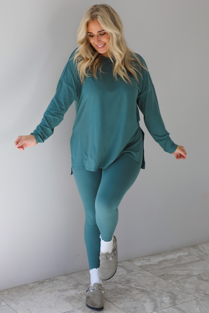 Woman wearing a teal two piece set that features an oversized long sleeved top and fitted leggings. The set is paired with high ankle socks and grey slide on shoes. The photo is taken indoors against a white wall with white marble flooring.