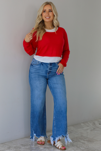 Woman wearing a red and white layered appearance top with dark wash denim jeans. The photo is taken indoors against a white wall with white marble flooring. 
