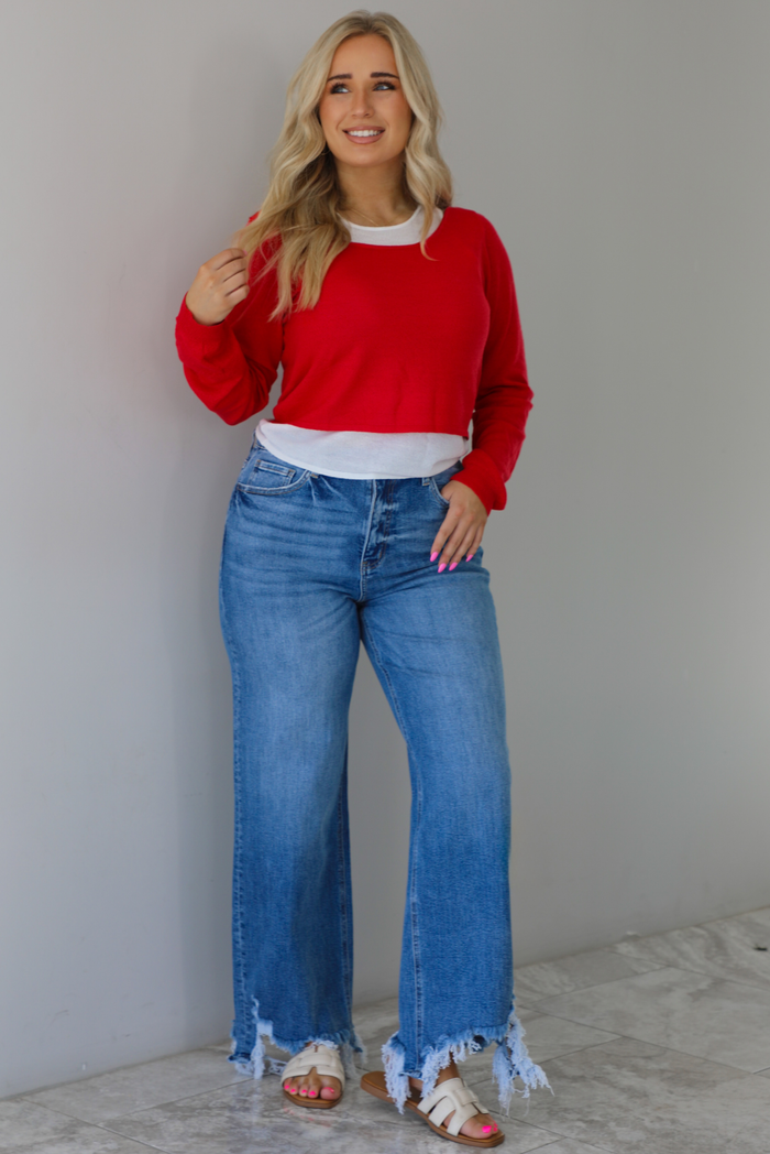 Woman wearing a red and white layered appearance top with dark wash denim jeans. The photo is taken indoors against a white wall with white marble flooring. 