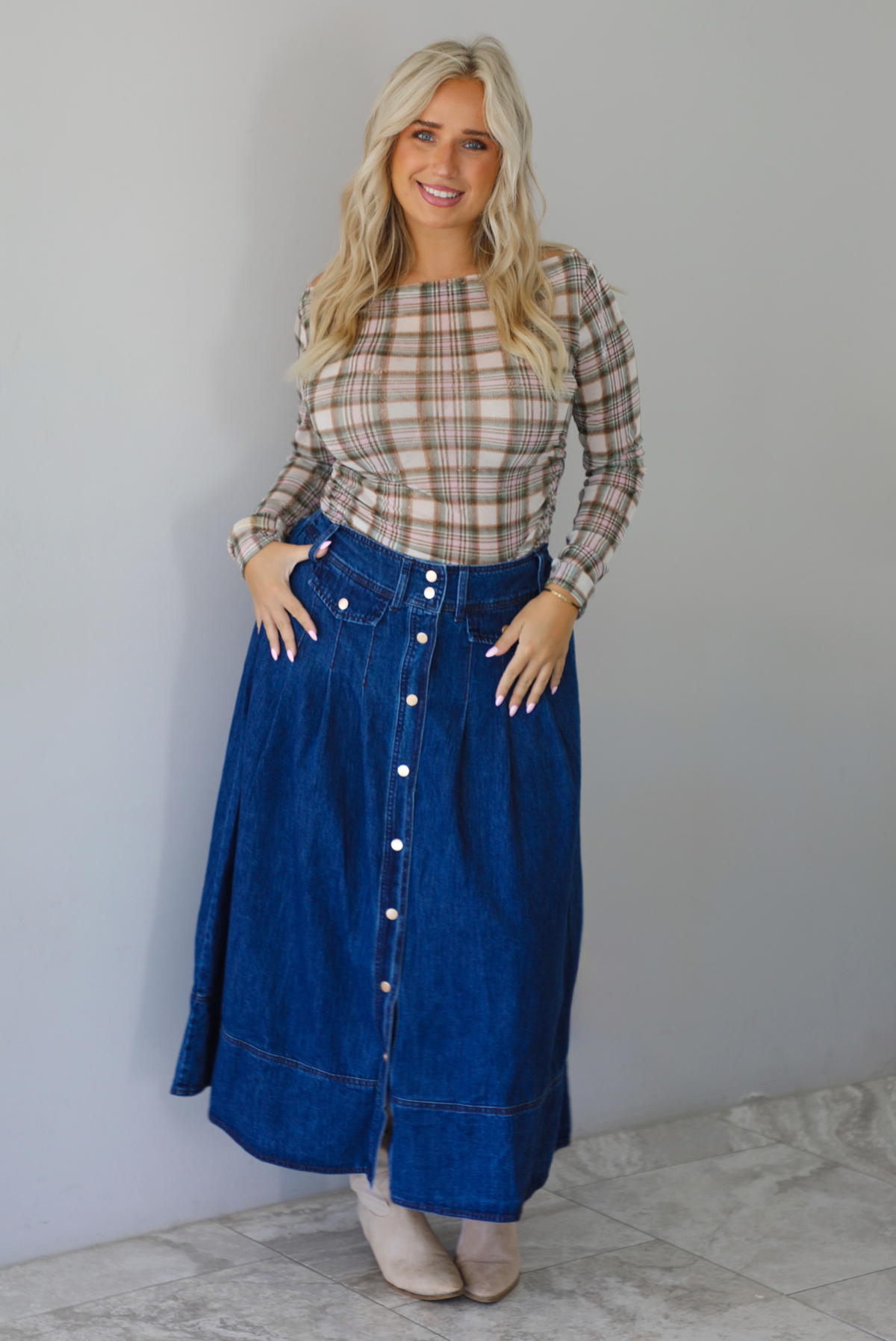 A woman wearing a beige and brown plaid long-sleeve top and a high-waisted blue denim button-front midi skirt, standing indoors against a white wall with white marble flooring. 