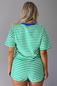 Woman wearing a green and off white striped pattern throughout that has blue trim details at the top of the neckline at the top and waistline on the shorts. The photo is taken indoors against a white wall and shows a closeup of the back of the set.