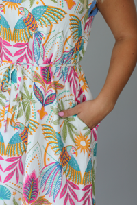 A woman wearing a short sleeved midi dress with a colorful tropical leaf print and light blue trim indoors against a white wall. The photo shows a closeup of the pocket on the side. 