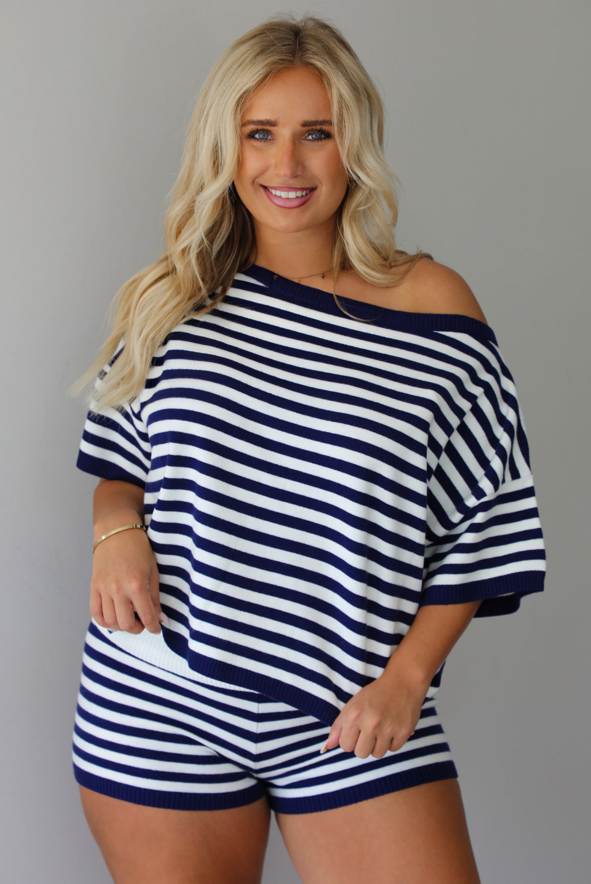 A woman wearing a navy and white horizontally striped off-the-shoulder top and matching striped shorts, standing indoors against a white wall.